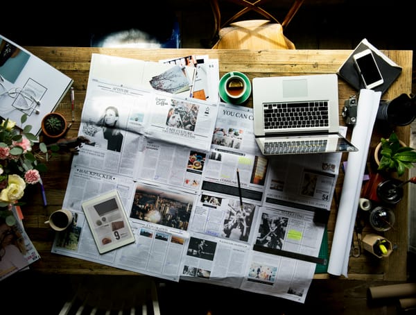A newspaper chaotically laid out on a desk with coffee, tablet, computer, phone, etc.